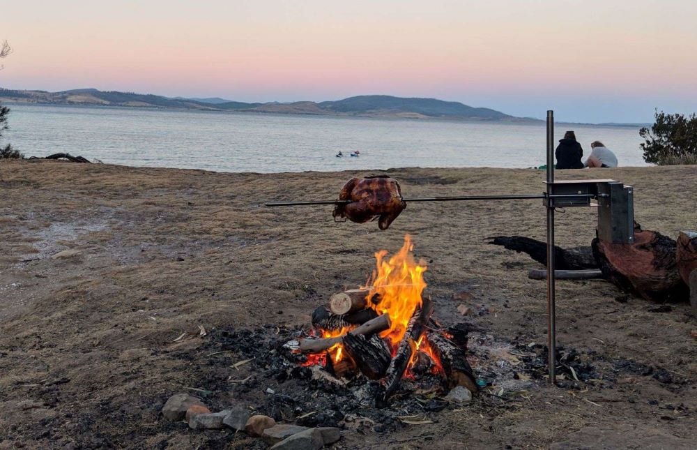 This is an image of the auspit stainless steel compact spit kit  cooking a chicken oven an open fire while out camping with the ocean in the background